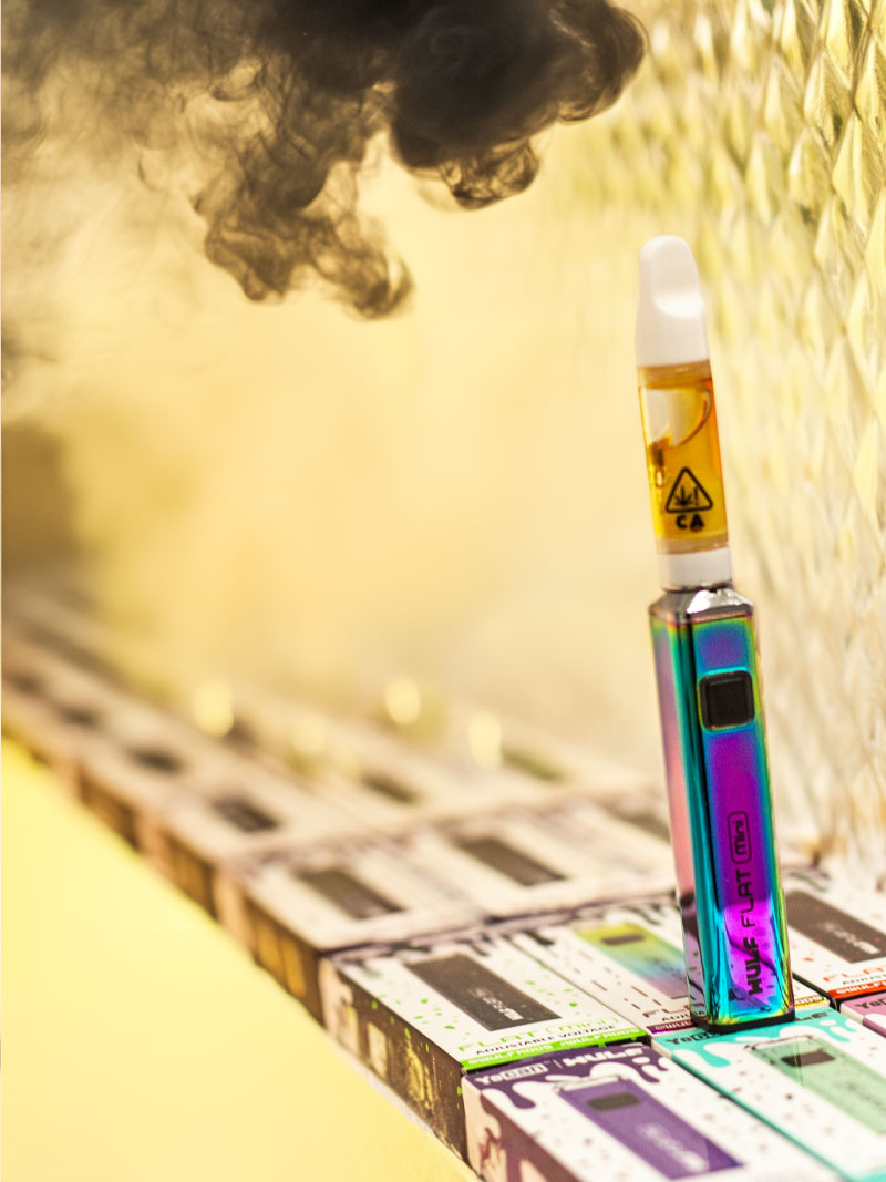 Wulf Flat Mini vaporizer resting on row of packaging near golden wall with thick smoke nearby